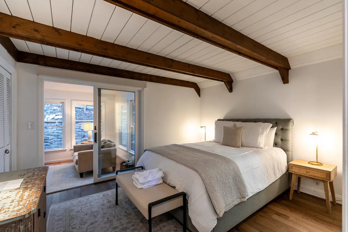 A tranquil queen bedroom featuring a plush bed positioned beside a large window, with a coffee table and lamp nearby. Natural light pours in, making the space feel restful and inviting—the ideal environment for relaxation and privacy.