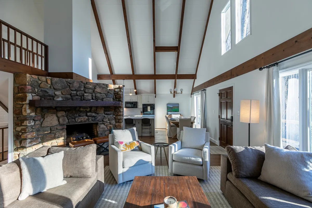A cozy portion of the living area centered on a stone fireplace, surrounded by comfortable seating, a coffee table, and warm wooden accents. Sunlight streams in, enhancing the inviting and relaxing ambiance ideal for unwinding or socializing by the fire.