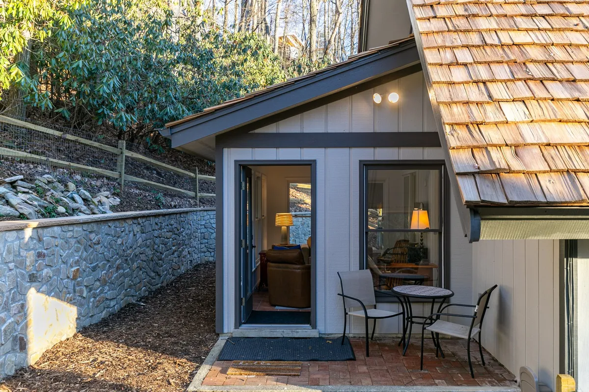 A patio with two chairs and a small table, overlooking the wooded landscape. The entrance to the living area is visible, bridging indoor comfort with outdoor enjoyment. The natural stone wall and tree backdrop add a sense of privacy and tranquility.