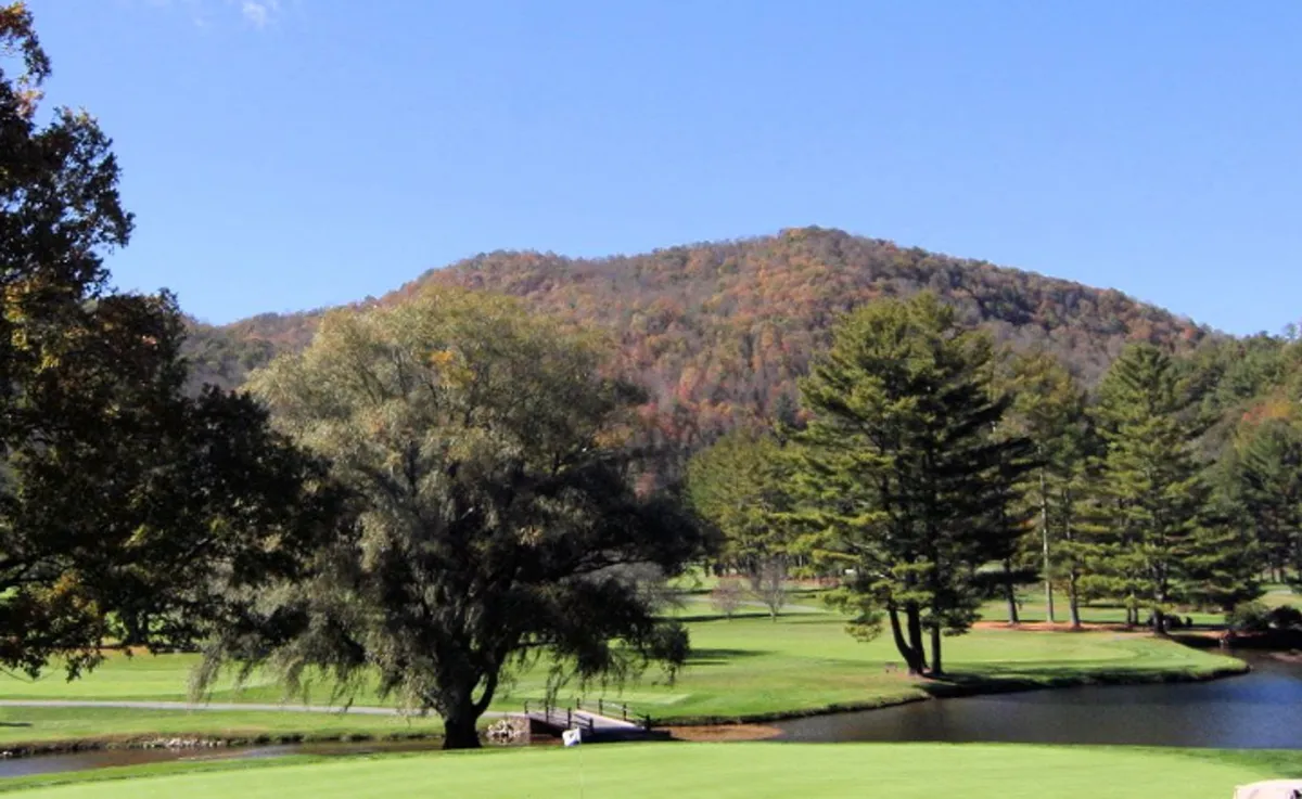 A sweeping view of a serene mountain landscape featuring a lush golf course, a peaceful lake, and clusters of trees, all framed by the striking backdrop of the surrounding mountains. The broad, open sky hints at the changing seasons and panoramic vistas unique to the High Country.