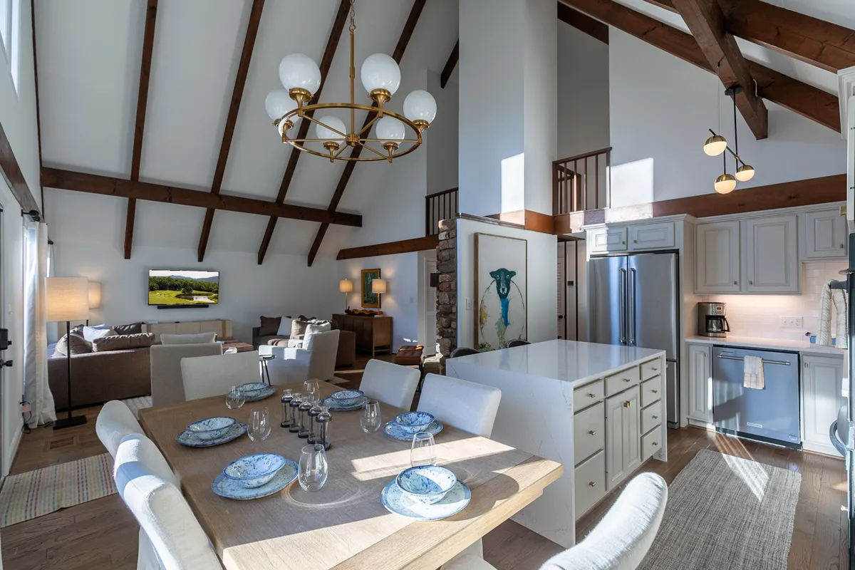 A welcoming angle featuring a dining table set with blue plates beneath a warm chandelier, the kitchen island in view, and part of the adjacent living area with cozy seating. The scene exudes comfort and readiness for shared meals or entertaining guests.
