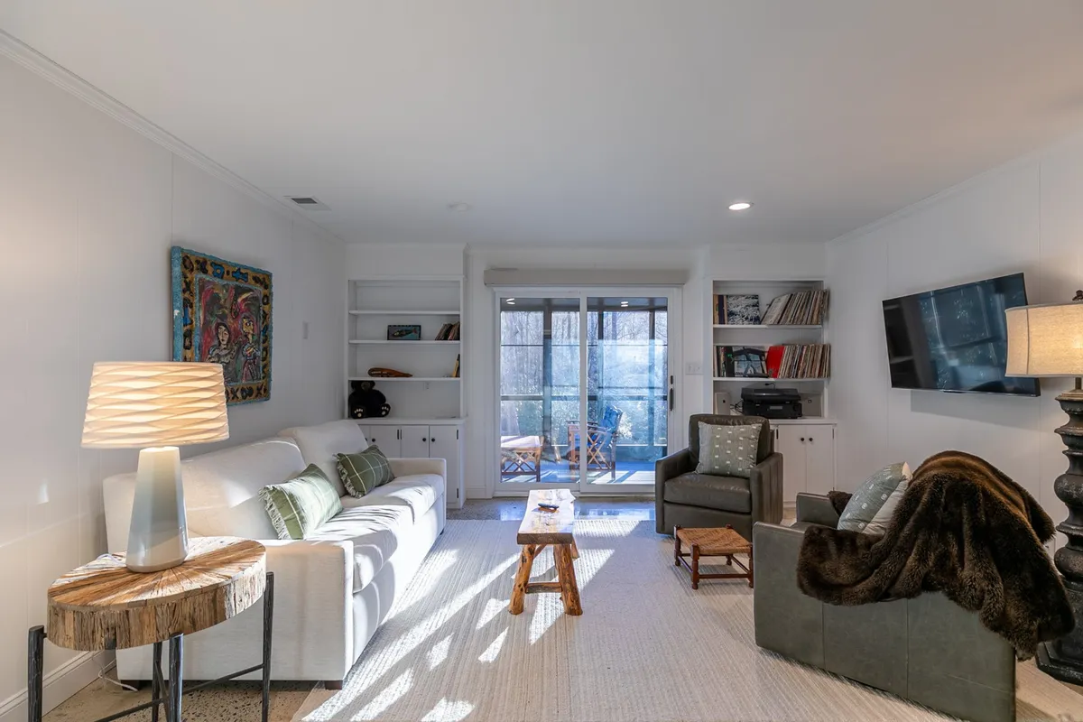 The scene shows a comfortable sofa positioned near a coffee table and lamp, with artwork above and a sliding glass door opening to a porch or patio. The soft lighting and layout invite both relaxation and socializing, while the glass door hints at easy access to outdoor spaces.