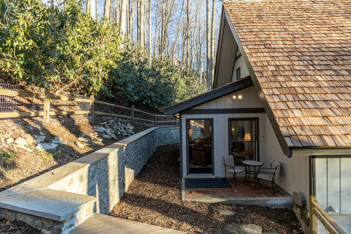 A welcoming entrance with a small table and chairs set on a stone patio, framed by wooden siding and a stone pathway. The scene suggests a peaceful transition from indoors to the vast, natural surroundings and hints at the secure, gated community setting.
