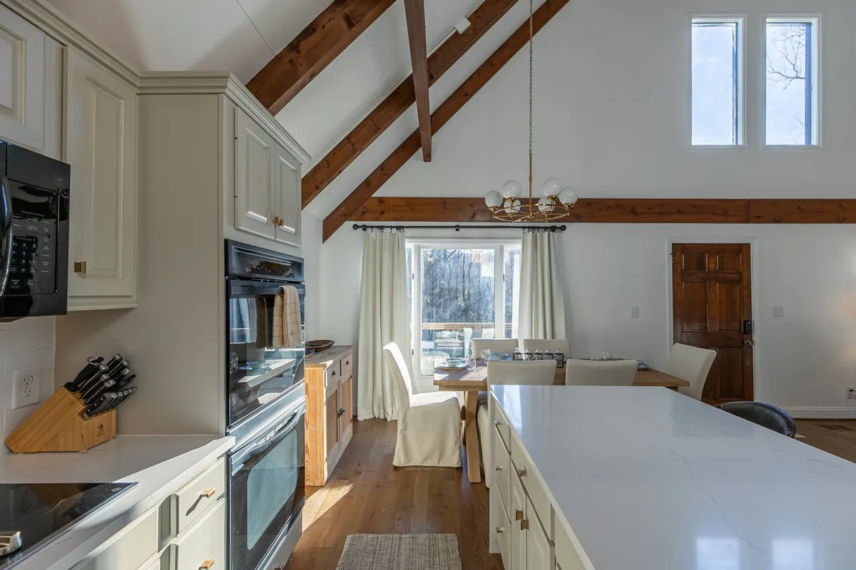 A wide view of the open-concept main living area, featuring a spacious kitchen island with granite countertops, a generous dining table, exposed wooden beams, and large windows that fill the space with natural light. The layout visually connects the kitchen, dining, and living spaces, emphasizing togetherness and effortless entertaining.