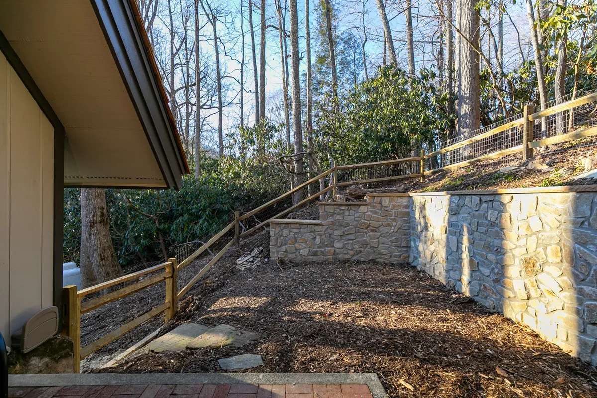 A landscaped exterior view featuring a stone retaining wall and wooden railing, nestled amidst natural mountain flora. The scene suggests peaceful walks and a secure, well-maintained environment.