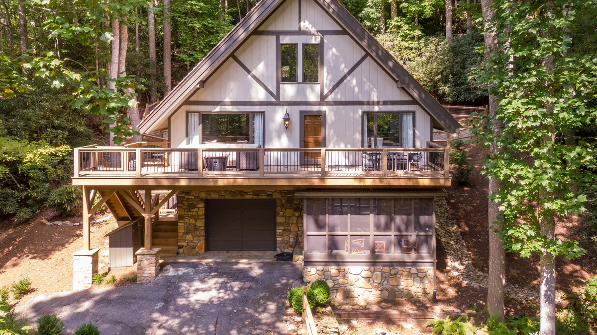 The image showcases a striking A-frame mountain home with a large stonework façade and spacious front deck, surrounded by lush greenery. The setting is tranquil, with ample space for relaxing on the deck, enjoying the views, and connecting with nature.