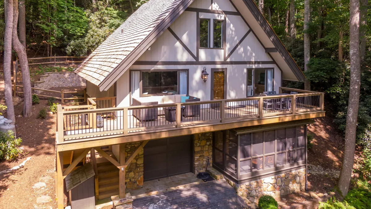 A welcoming exterior view of the A-frame architecture, featuring a spacious wooden deck and stone accents. The setting suggests an inviting outdoor space ideal for relaxing or spending time with family and pets, surrounded by a serene mountain backdrop.