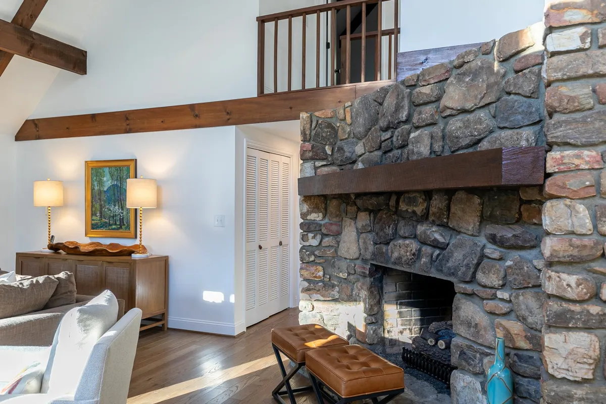 This cozy interior image features a stone fireplace framed by wooden beams, a plush sofa, wall art, and soft table lamps. The setting feels intimate and warm, emphasizing a space perfect for gathering, reading, or relaxing after a day of adventure.