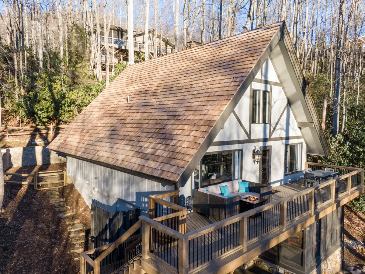 This image features the signature A-frame architecture of the house, a spacious wooden deck, and a cozy outdoor seating area. The deck overlooks lush mountain surroundings, and the scene evokes moments of sipping coffee at sunrise or gathering with friends for evening bonfires and stargazing.